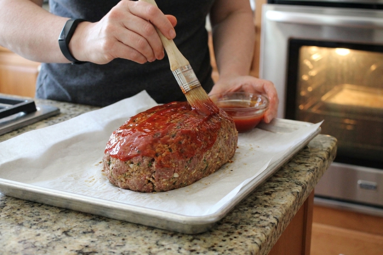 A pastry brush spreading ketchup glaze over a partially baked meatloaf on a sheet pan near an oven