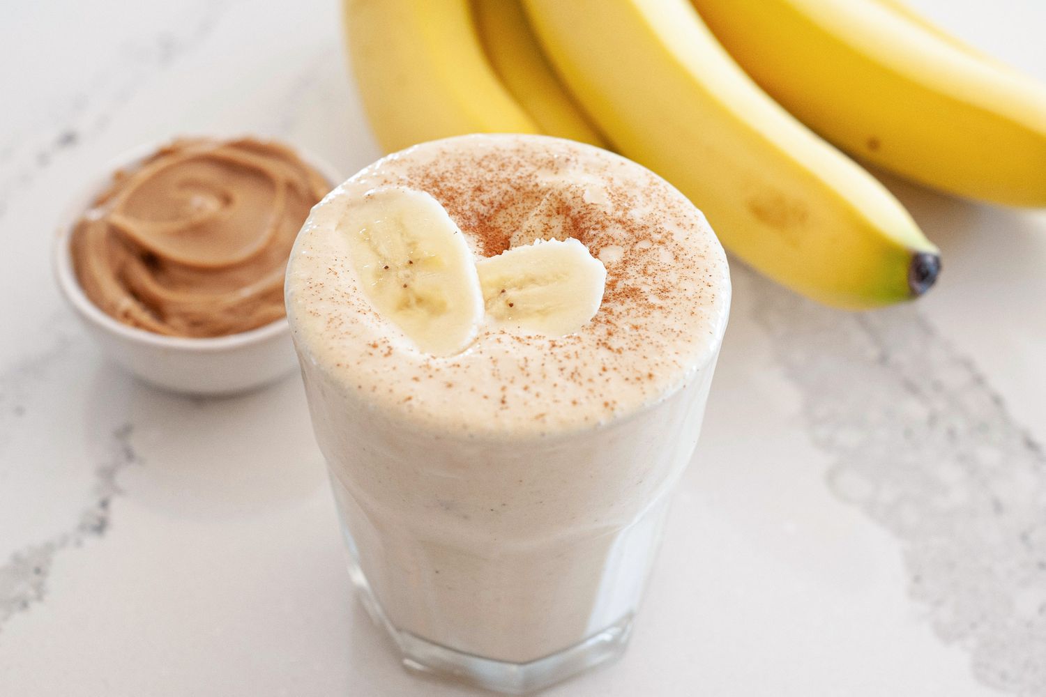 A peeled banana, a spoonful of peanut butter, a small pitcher of milk, vanilla extract, and ice cubes arranged on a tidy kitchen counter