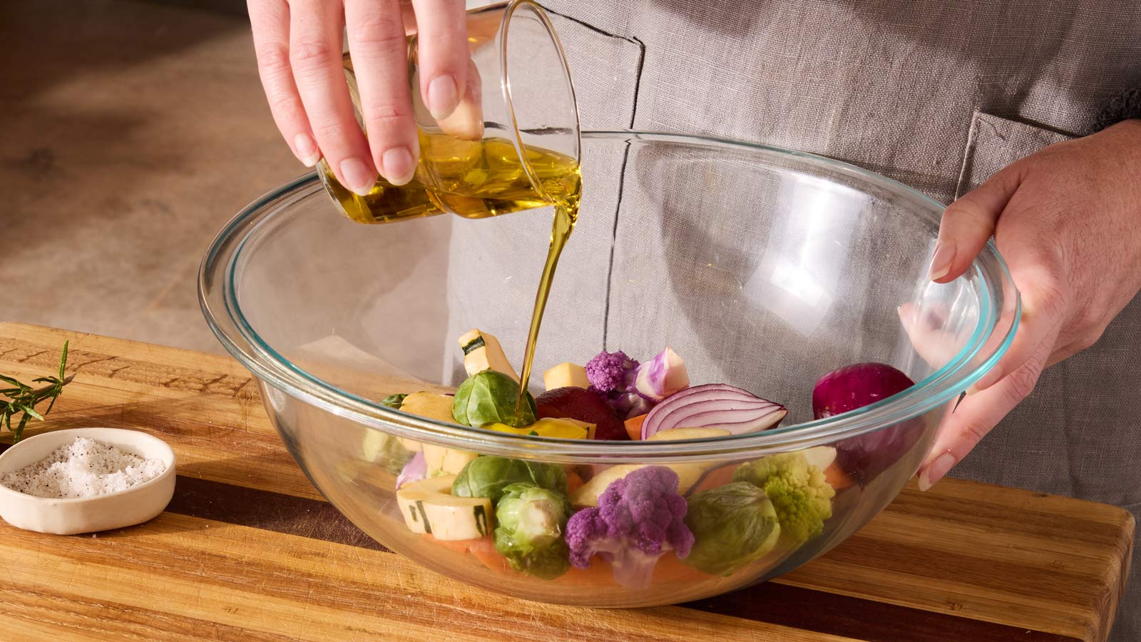 A person tossing chopped vegetables with olive oil and seasonings in a large stainless steel mixing bowl on a kitchen counter