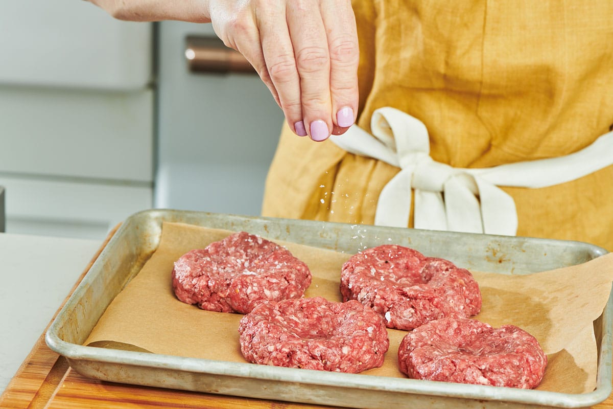 A person’s hands gently shaping raw ground beef into burger patties on parchment paper on a kitchen counter