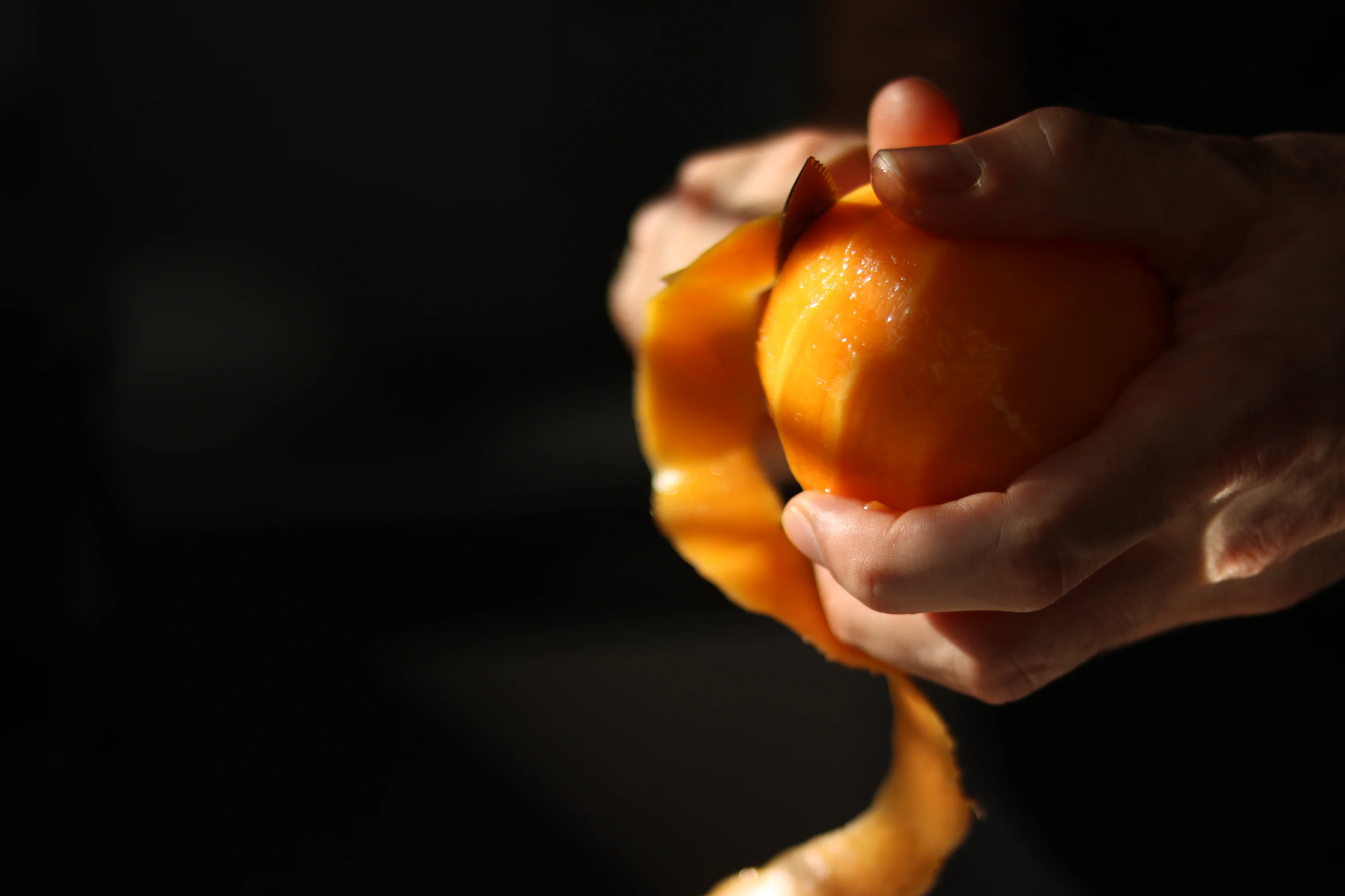 A person’s hands using a vegetable peeler to cut a wide strip of orange peel over a wooden cutting board on a kitchen counter