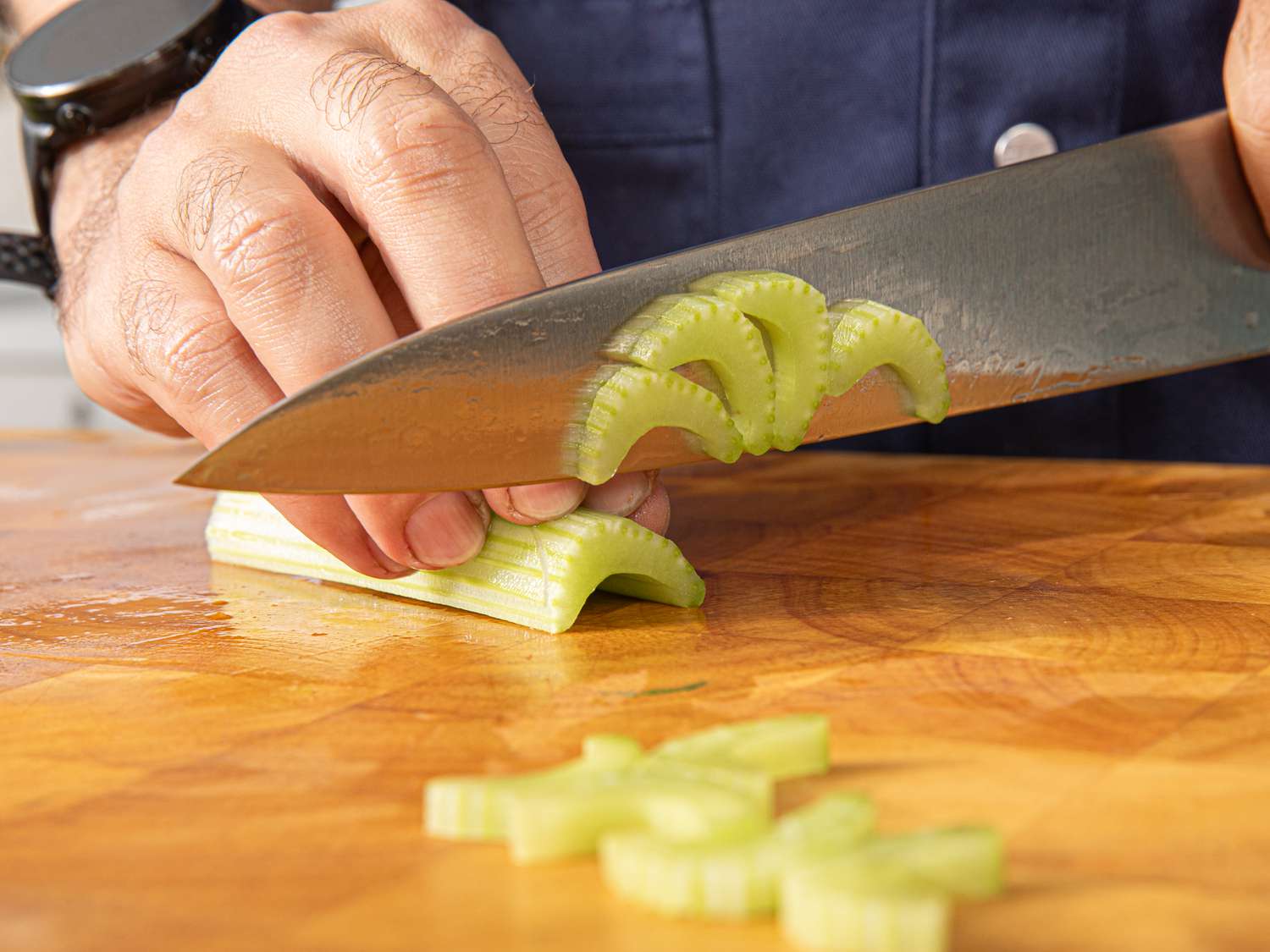 A pile of chopped celery with a few celery leaves on a wooden cutting board next to a chef's knife, bright kitchen counter lighting