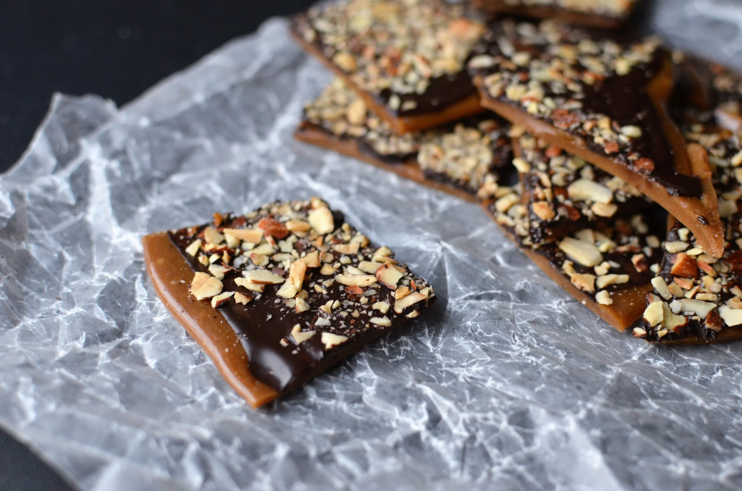 A pile of homemade English toffee pieces with a glossy chocolate top and chopped toasted almonds on a parchment-lined baking sheet in a warm home kitchen