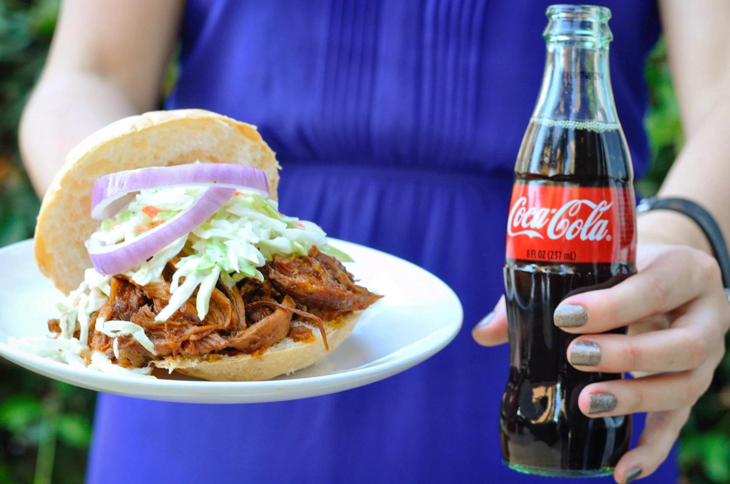 A piled-high pulled pork sandwich on a toasted bun with glossy cola barbecue sauce, pickles, and a simple slaw on a wooden table