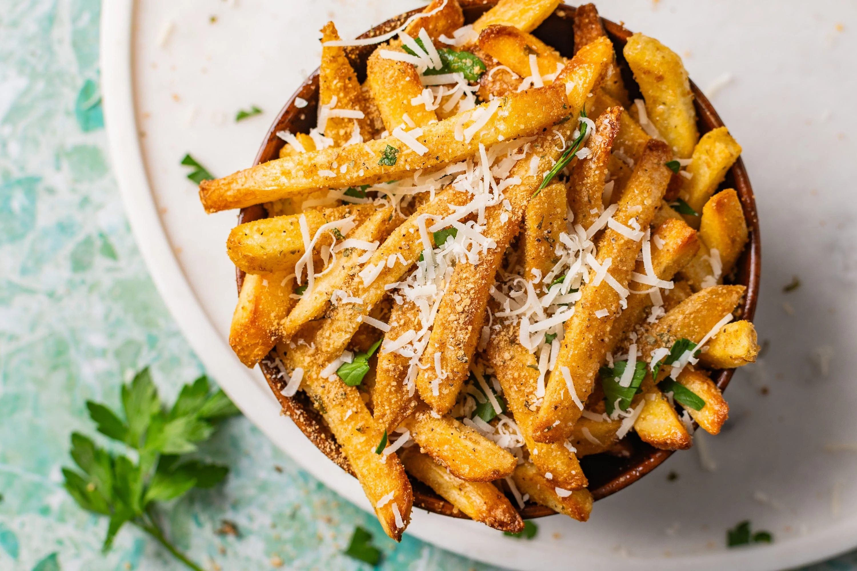 A piled platter of crispy golden fries tossed with Parmesan and parsley, with a small bowl of creamy garlic aioli on a warm wooden table