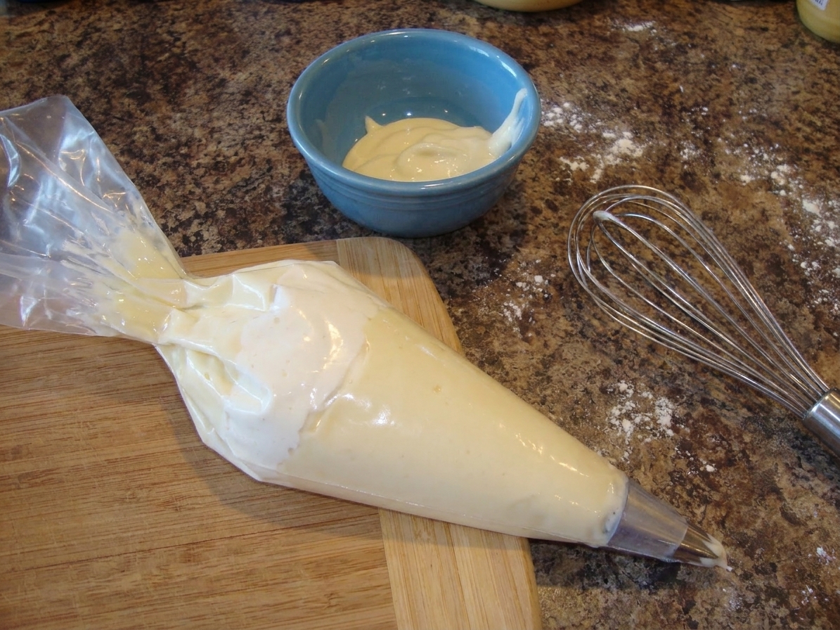 A piping bag filled with pale vanilla gel resting on a cutting board beside a small bowl and whisk on a kitchen counter