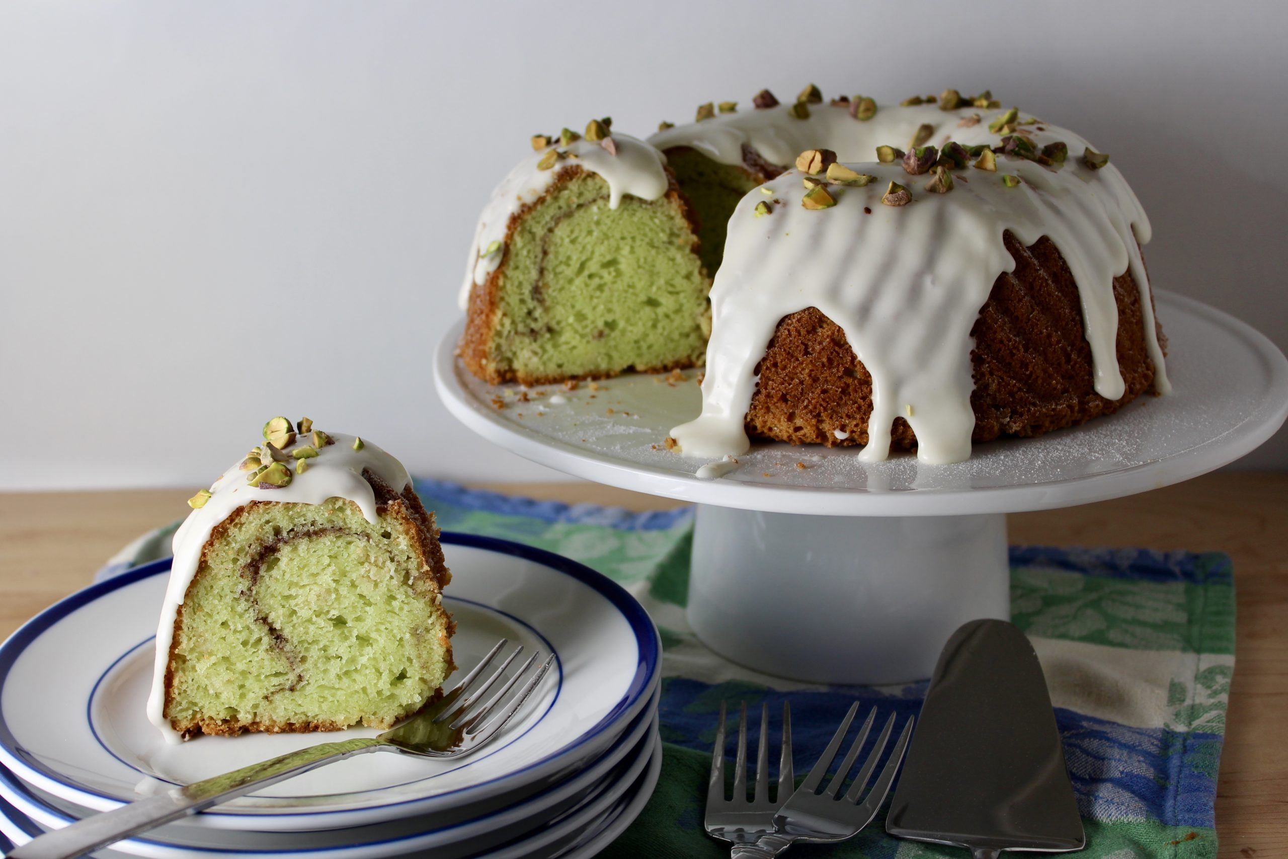 A pistachio bundt cake on a cooling rack while a pale green glaze is being drizzled over the top from a spoon, creating glossy drips down the sides