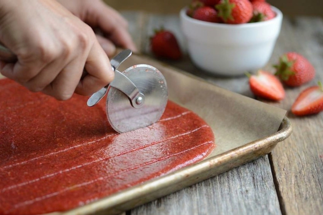 A pizza cutter slicing a sheet of strawberry fruit leather into even strips on parchment paper