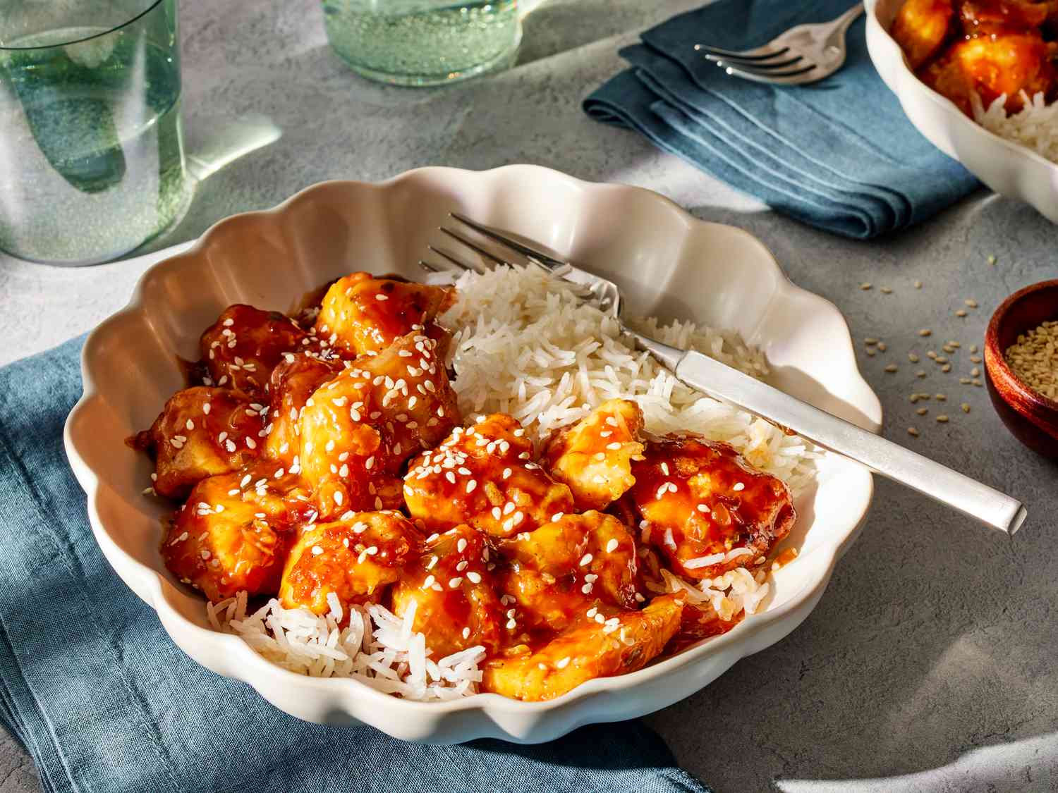 A plate of crispy General Tso chicken coated in a glossy dark amber sauce with sliced scallions and sesame seeds on a simple white plate on a warm kitchen table