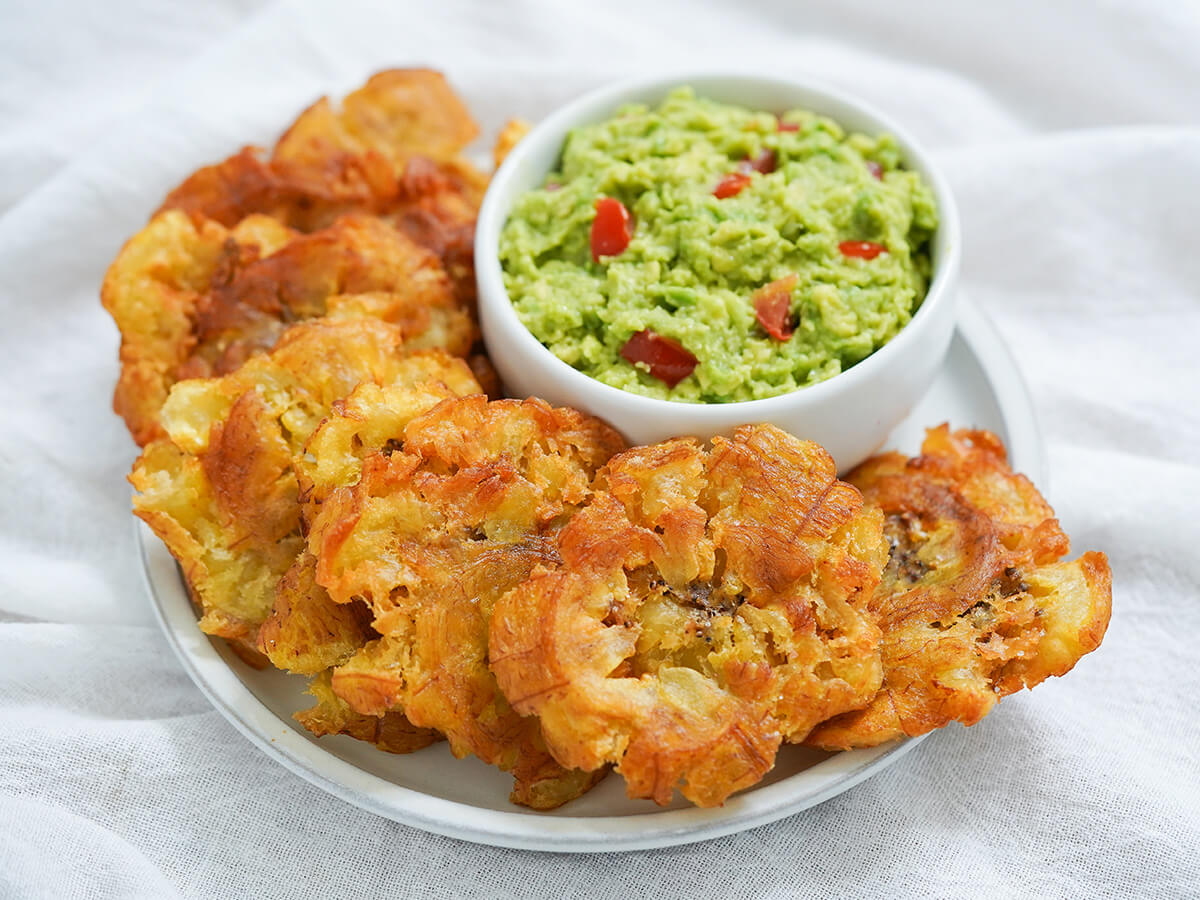 A plate of crispy golden tostones stacked with a small bowl of garlicky dipping sauce on a warm wooden table in a bright home kitchen
