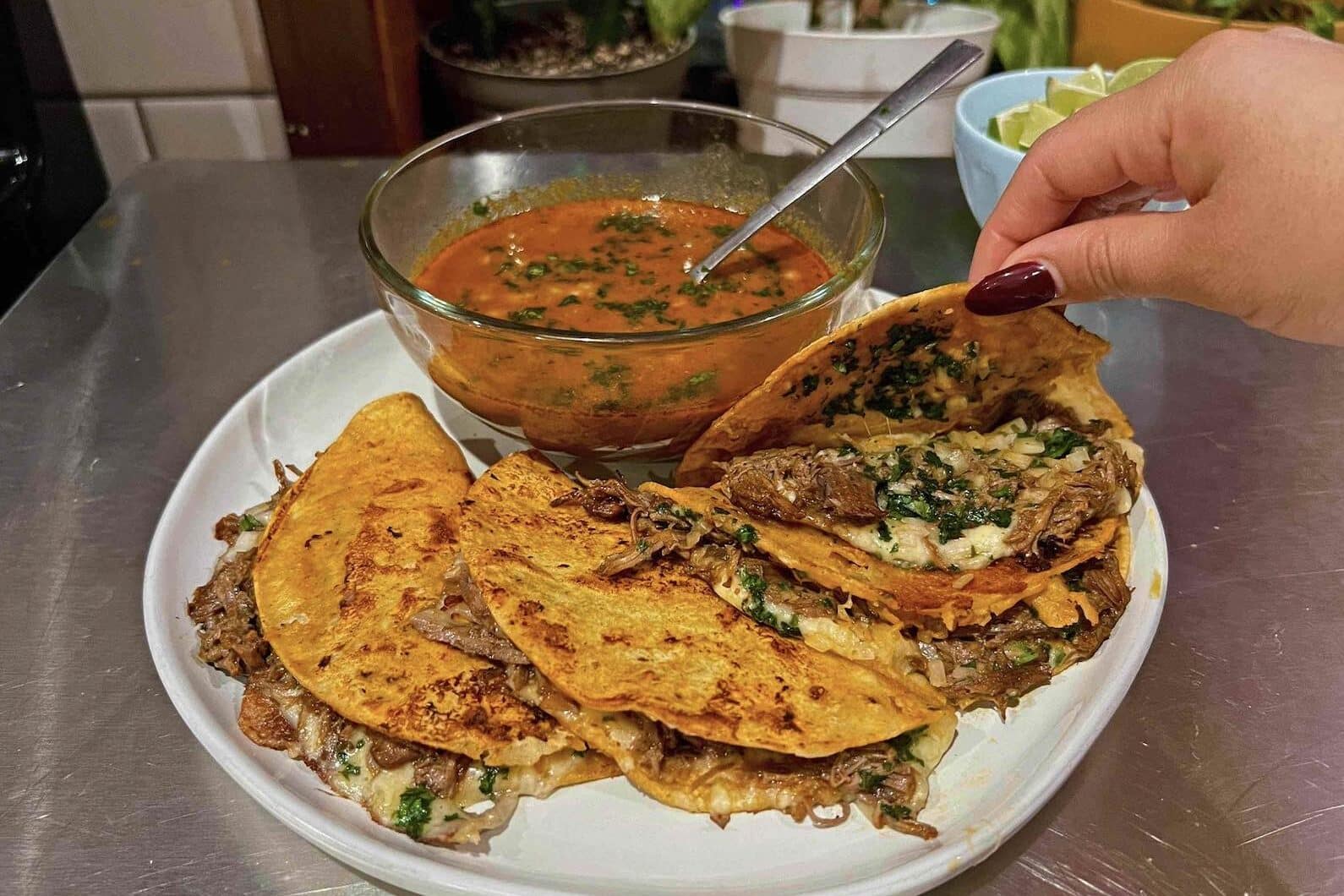 A plate of crispy quesabirria tacos folded and browned until crisp, served with a small bowl of deep red birria broth for dipping, topped with chopped onion and cilantro on a warm kitchen table