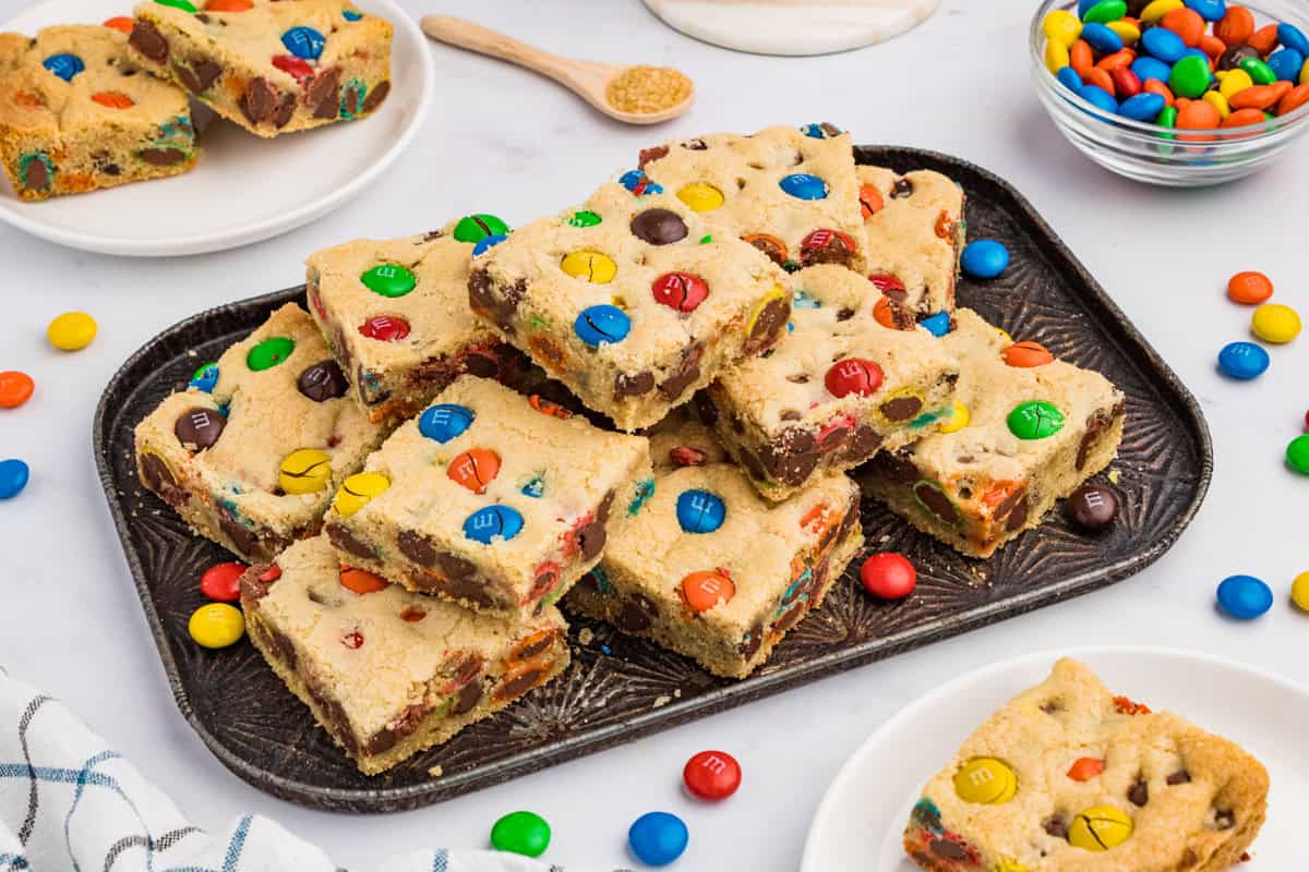 A plate of freshly baked M&M cookies with crisp golden edges and colorful candies, set on a simple wooden kitchen table in bright natural window light