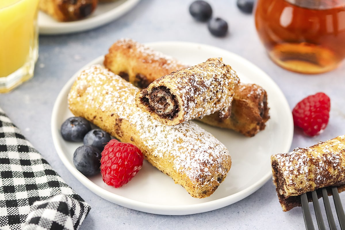 A plate of golden French toast roll-ups coated in cinnamon sugar, sliced to show a gooey Nutella center with banana, on a simple breakfast table with a mug of coffee