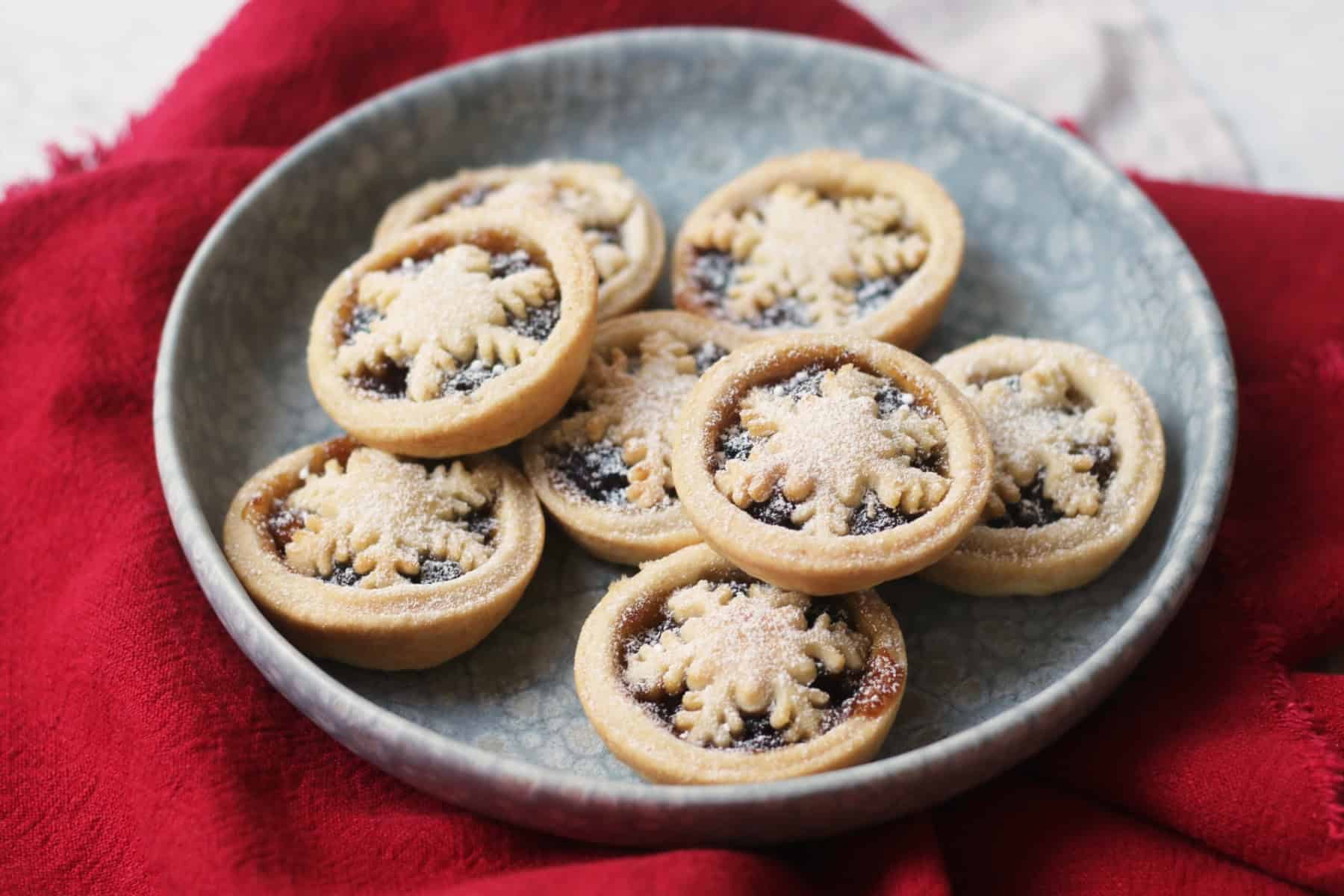 A plate of golden-brown mince pies with crisp pastry tops, lightly dusted with powdered sugar on a wooden table under warm kitchen lighting
