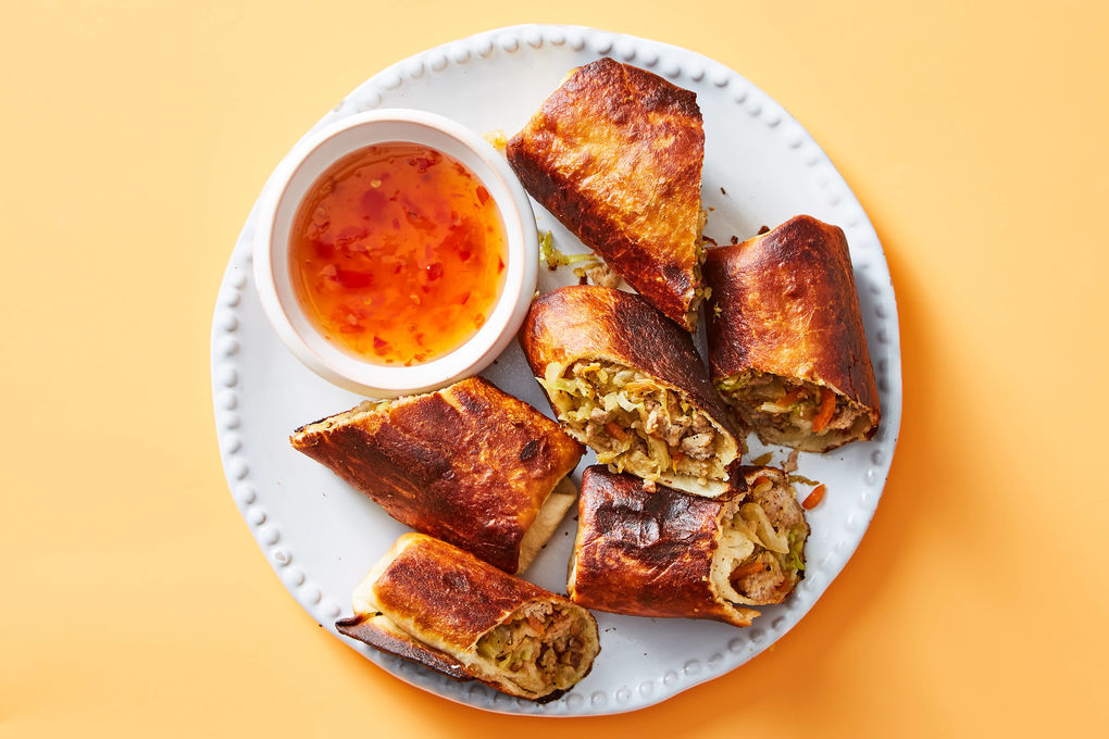 A plate of golden brown pork egg rolls sliced open to show the pork and cabbage filling, served with a small bowl of sweet chili sauce on a wooden table
