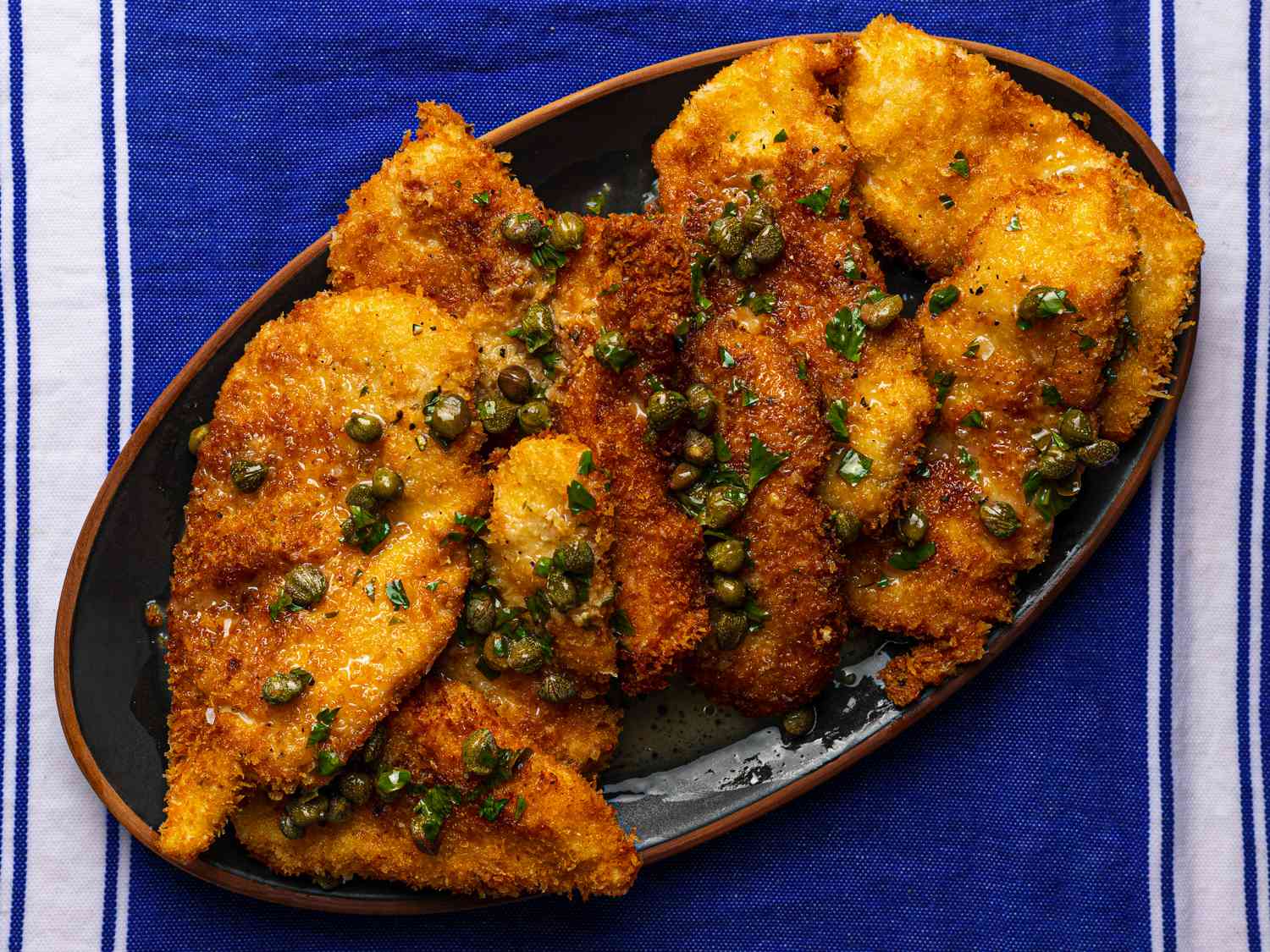 A plate of golden crispy chicken cutlets topped with glossy lemon caper butter sauce on a warm dinner table, with a skillet in the background
