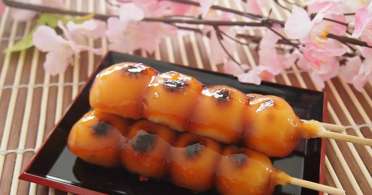 A plate of mitarashi dango skewers coated in a glossy sweet soy glaze, with a small bowl of extra sauce on a warm kitchen table