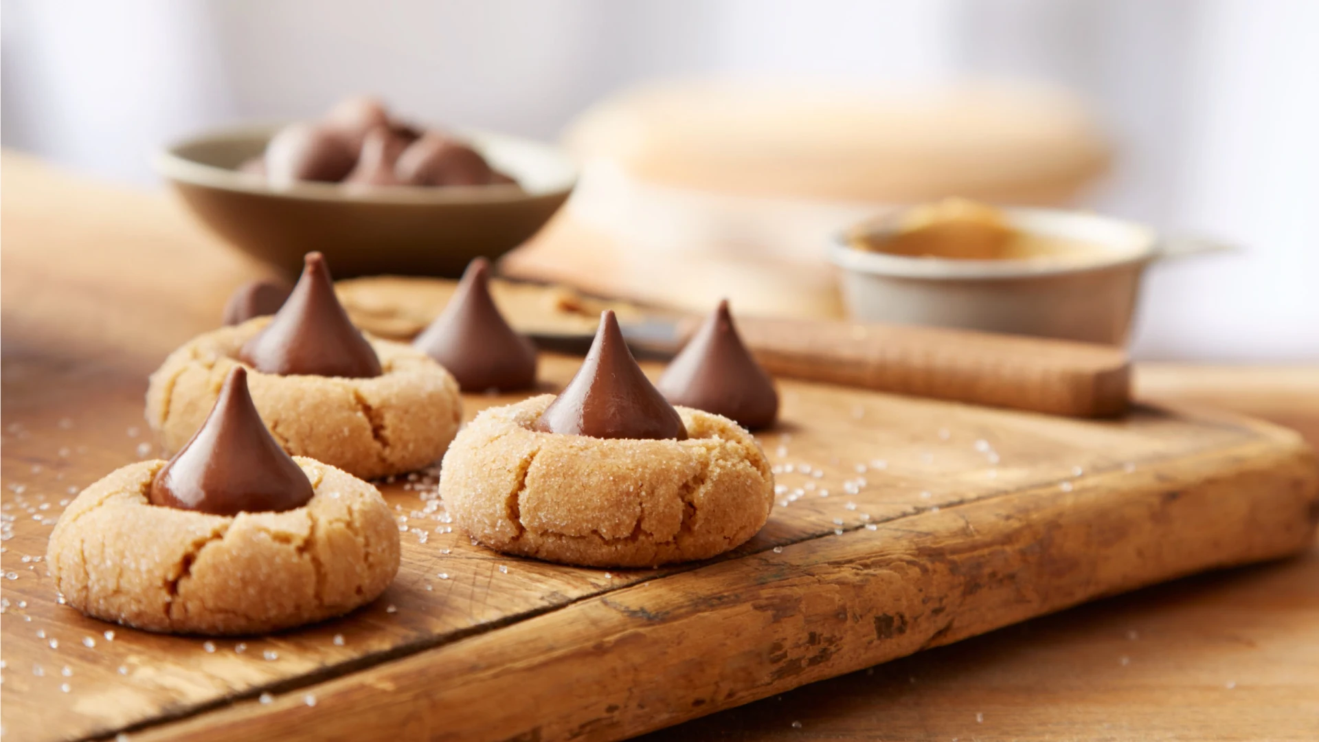 A plate of peanut blossom cookies with chocolate kisses pressed into the centers, on a warm wood table with soft kitchen lighting