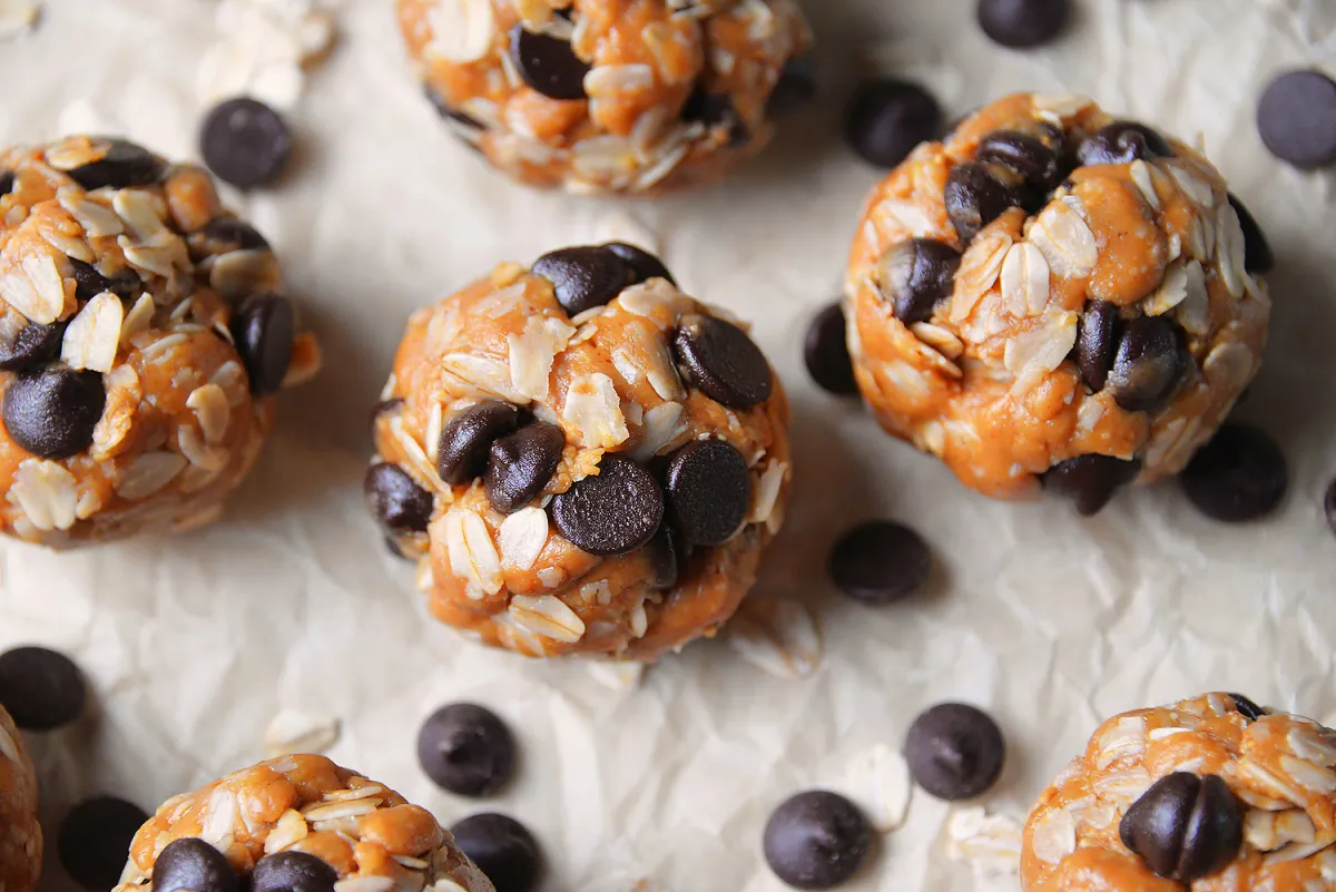 A plate of peanut butter oat energy bites with mini chocolate chips on a light kitchen counter, with a small bowl of oats and a jar of peanut butter nearby