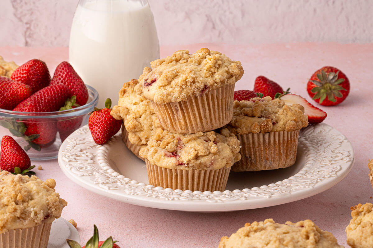 A plate of tall, golden strawberry muffins with visible strawberry pieces, set on a light kitchen table with a soft linen napkin and morning window light