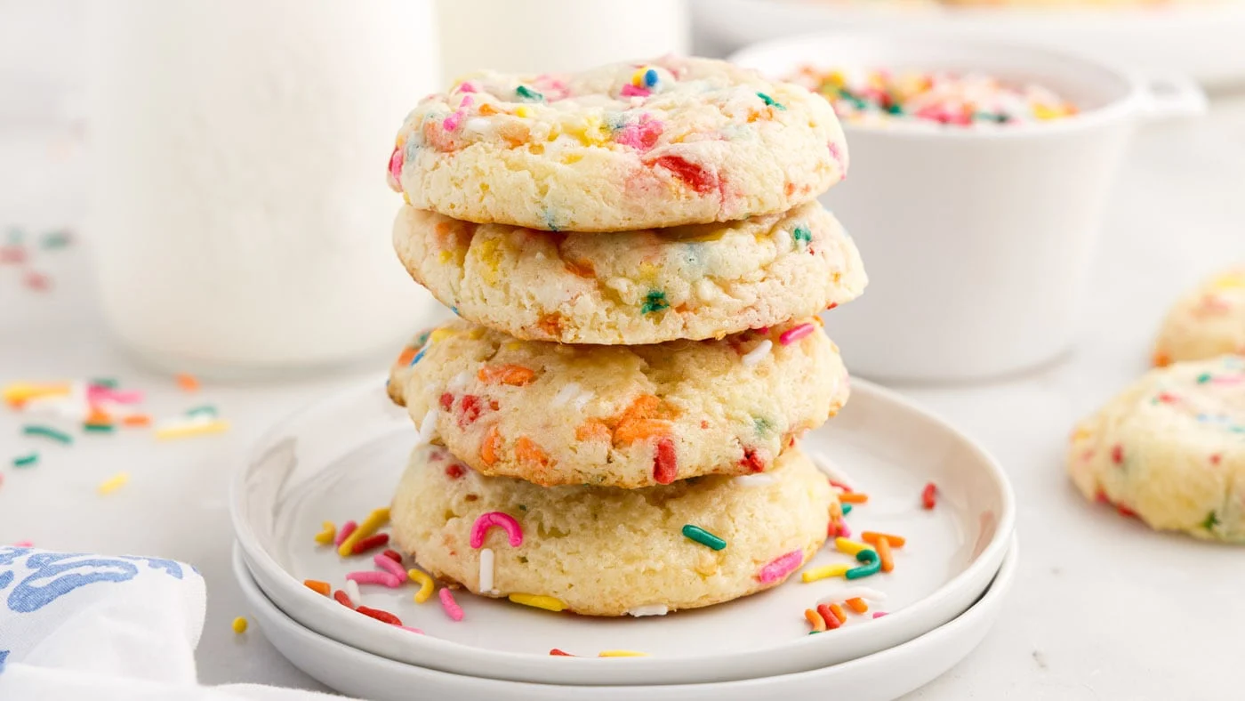 A plate of thick funfetti cake mix cookies with rainbow sprinkles, soft centers, and lightly crisp edges on a wooden kitchen table with warm natural light