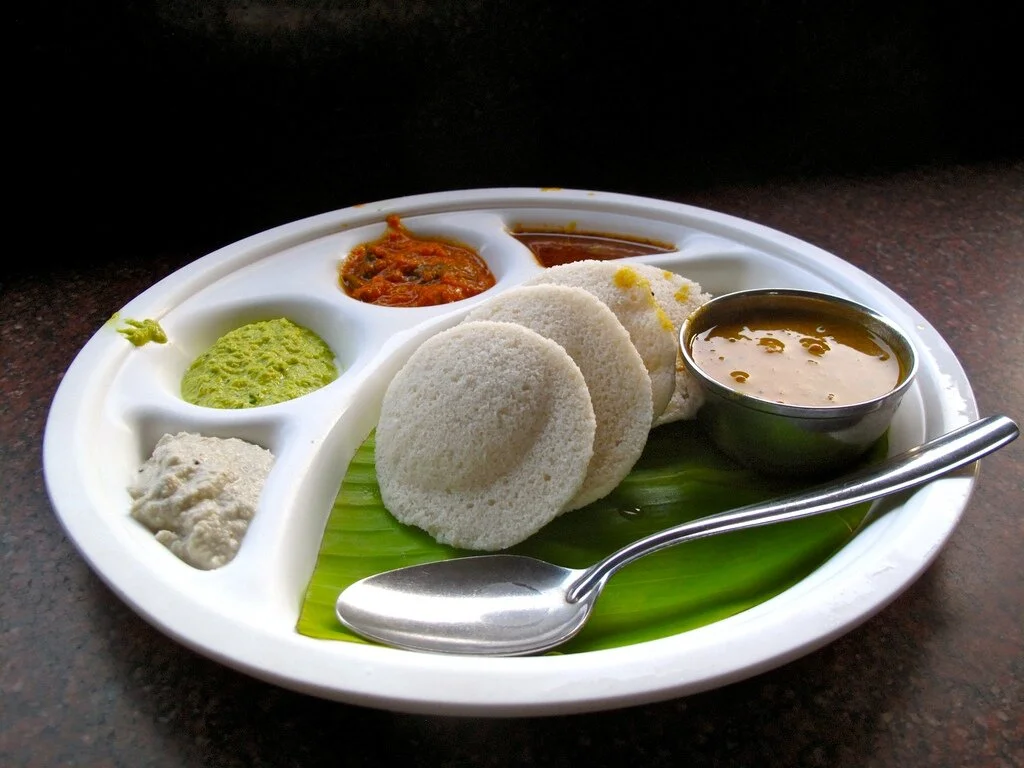 A plate of warm, fluffy idli stacked with a bowl of coconut chutney and a small bowl of sambar on a wooden table in a bright home kitchen