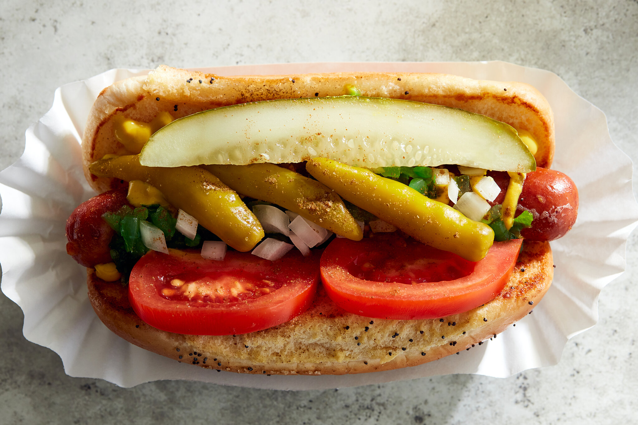 A plated Chicago-style hot dog in a poppy seed bun topped with yellow mustard, bright green relish, chopped onions, tomato wedges, a pickle spear, sport peppers, and a dusting of celery salt on a simple kitchen table