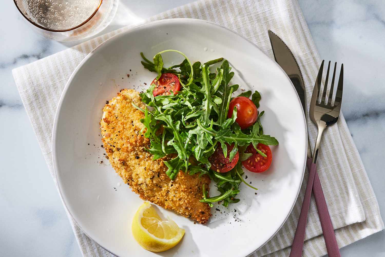 A plated Chicken Milanese cutlet with a lemony arugula salad on top, golden crust visible, on a simple white plate at a family dinner table