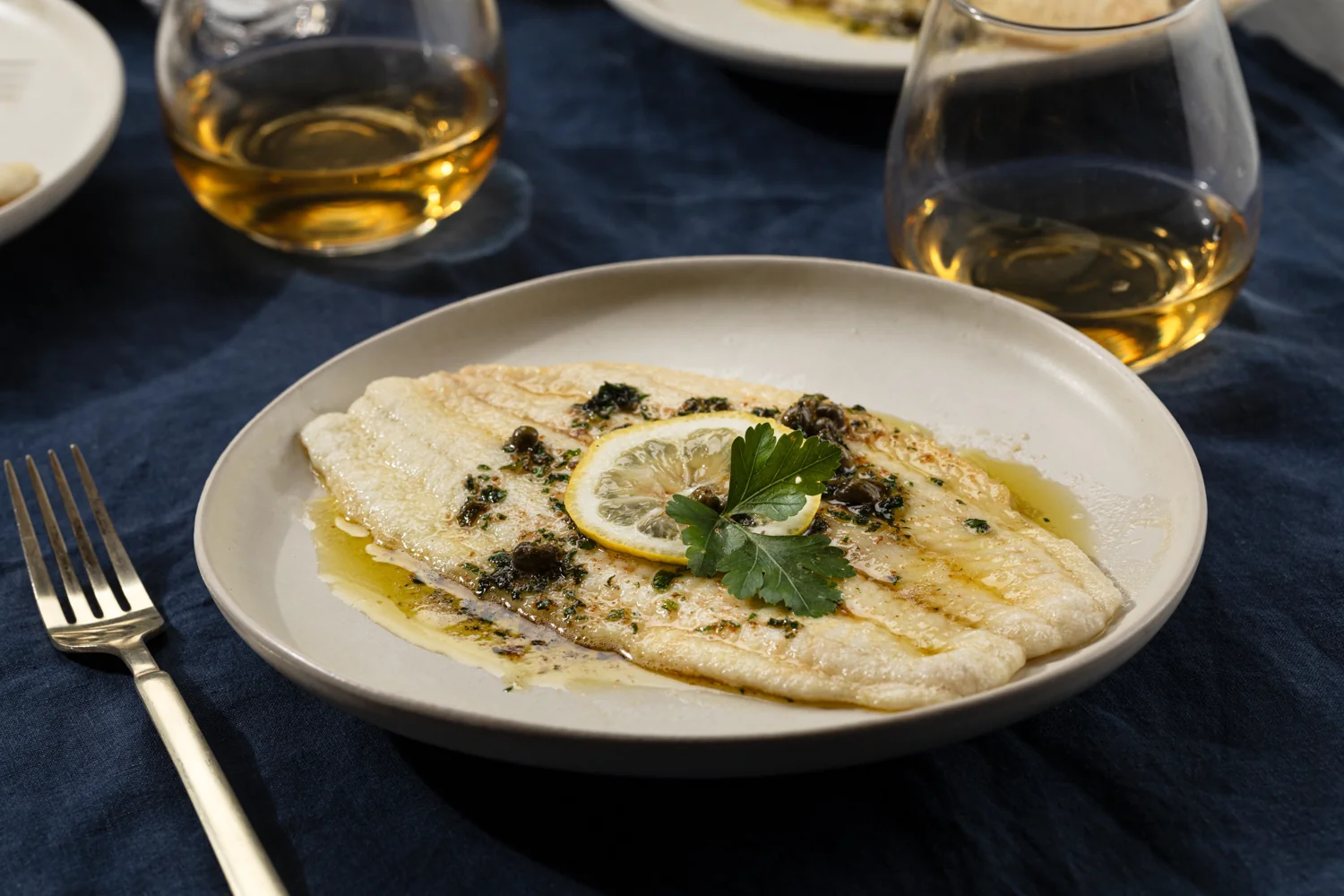 A plated Dover sole meunière on a white plate with browned butter lemon caper sauce, chopped parsley, and a lemon wedge, set on a warm-toned kitchen table