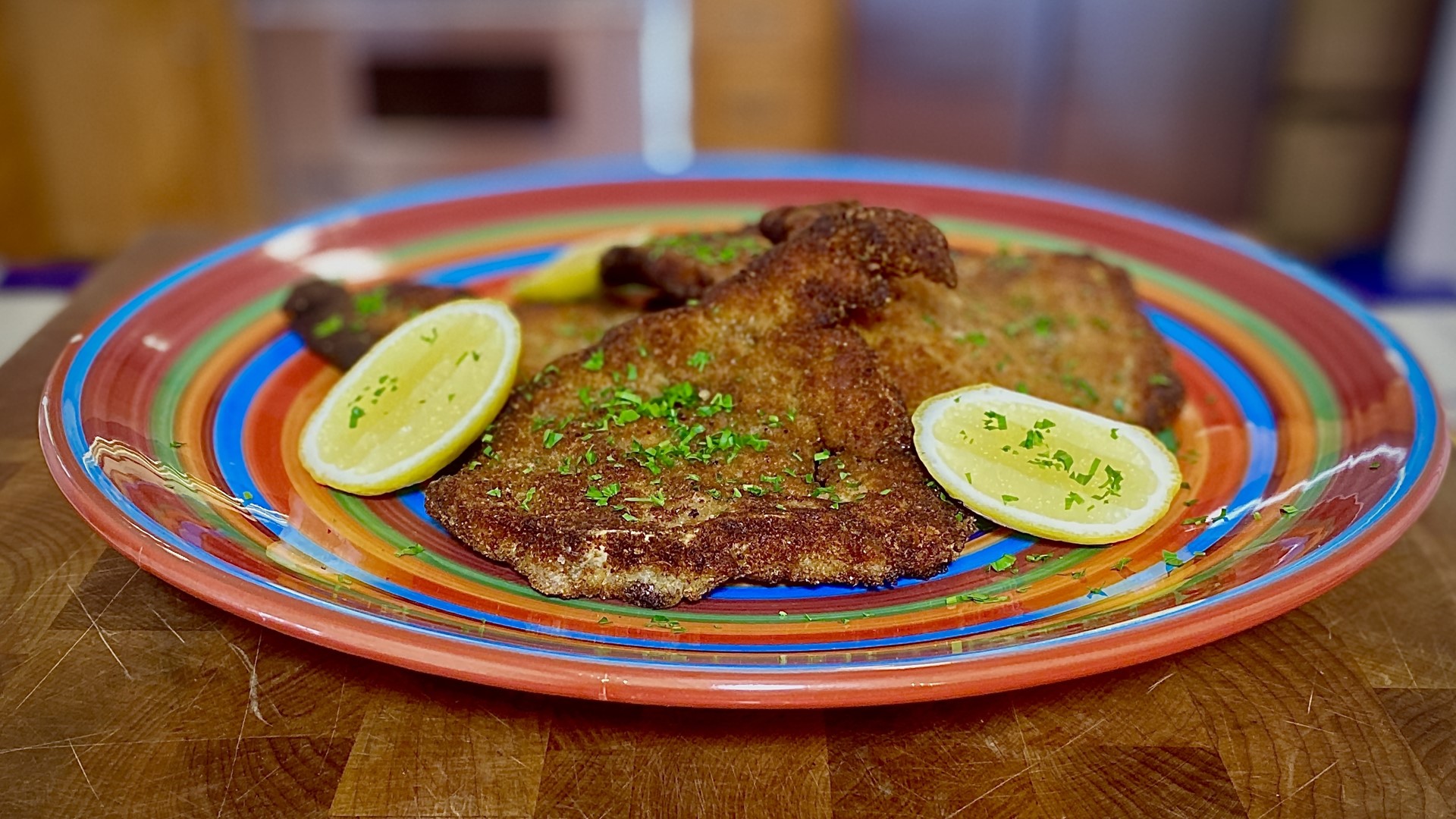 A plated German schnitzel with a crisp golden crust, lemon wedges, and chopped parsley on a simple white plate on a warm kitchen table