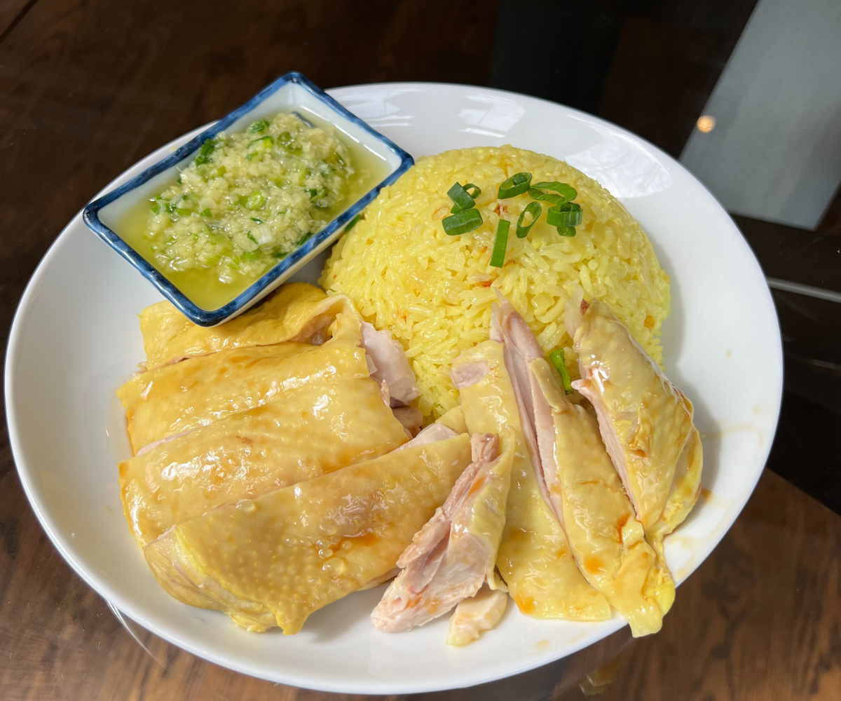 A plated Hainanese chicken rice set with sliced poached chicken, a mound of fragrant rice, cucumber slices, a small bowl of clear broth, and two dipping sauces on a warm kitchen table