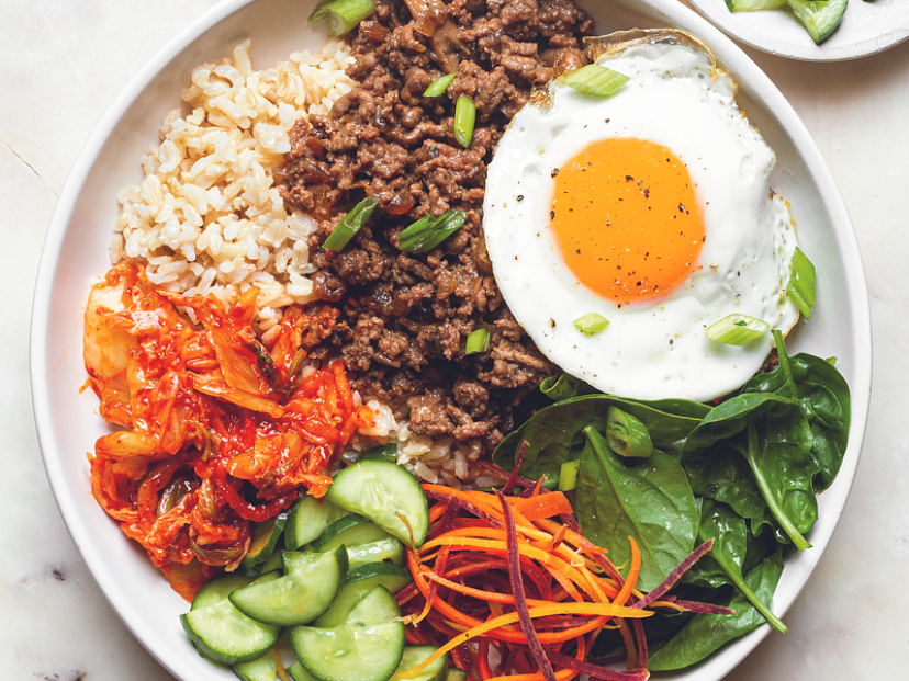A plated Korean beef bowl with glossy ground beef over white rice, topped with sliced green onions and sesame seeds on a simple wooden table setting