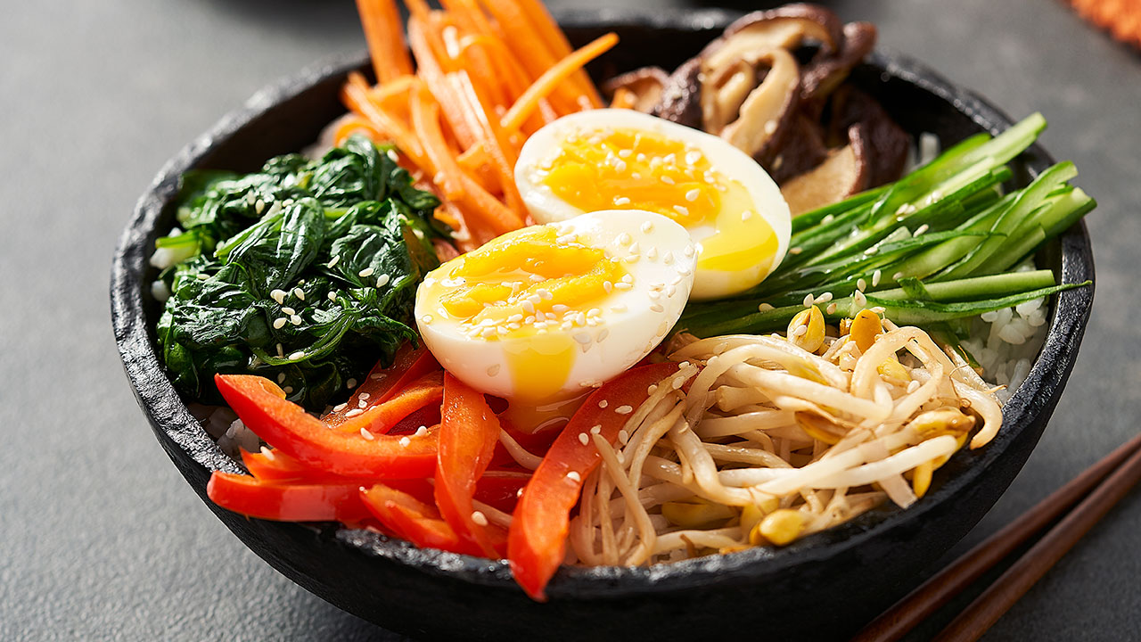 A plated Korean bibimbap bowl with white rice, sautéed spinach, carrots, zucchini, mushrooms, seasoned ground beef, a sunny-side-up egg, and a spoonful of gochujang sauce on a wooden table