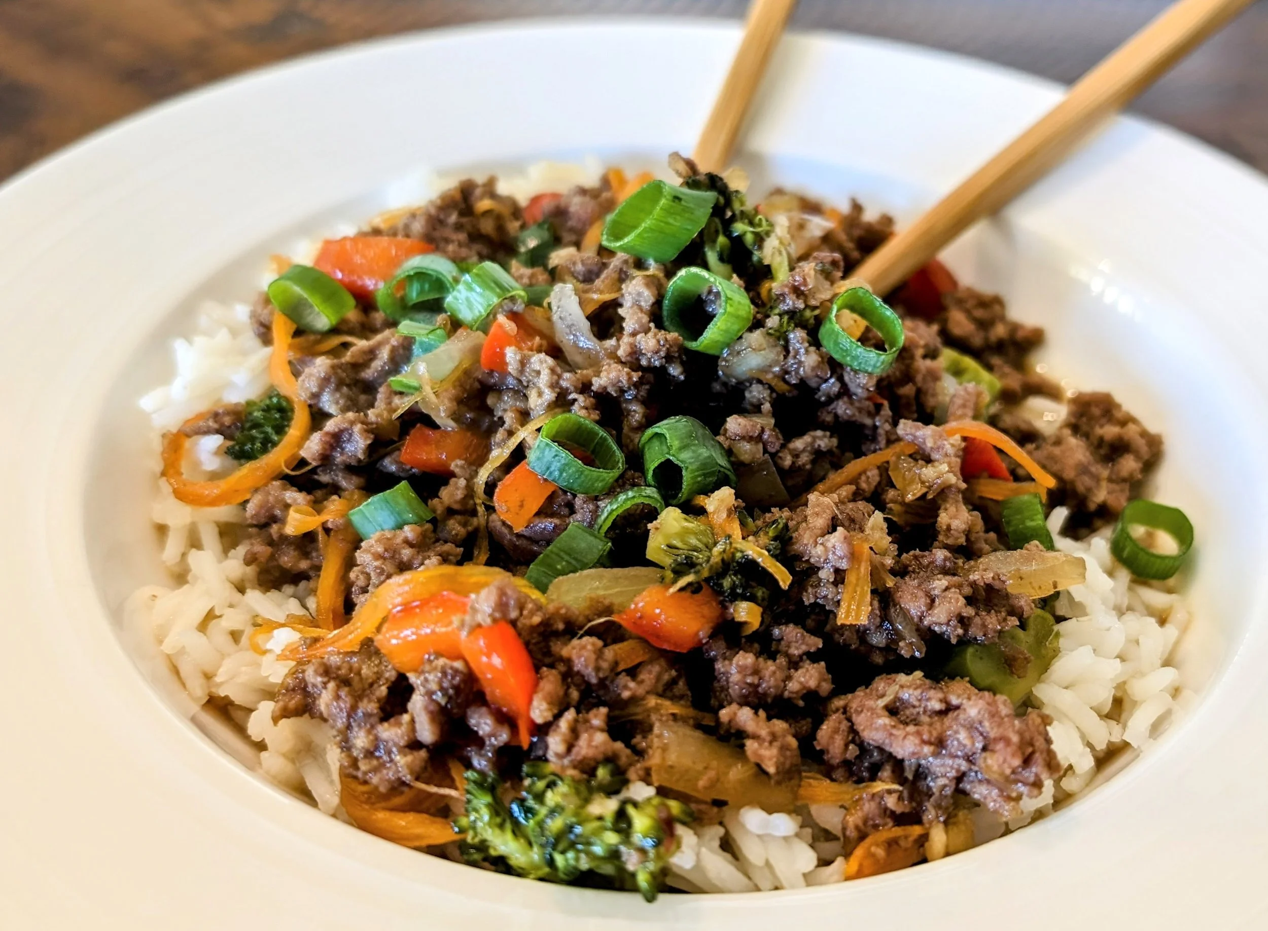 A plated Korean-inspired ground beef bowl with glossy soy-garlic beef over white rice, sliced cucumbers, shredded carrots, and sesame seeds on a light wood table
