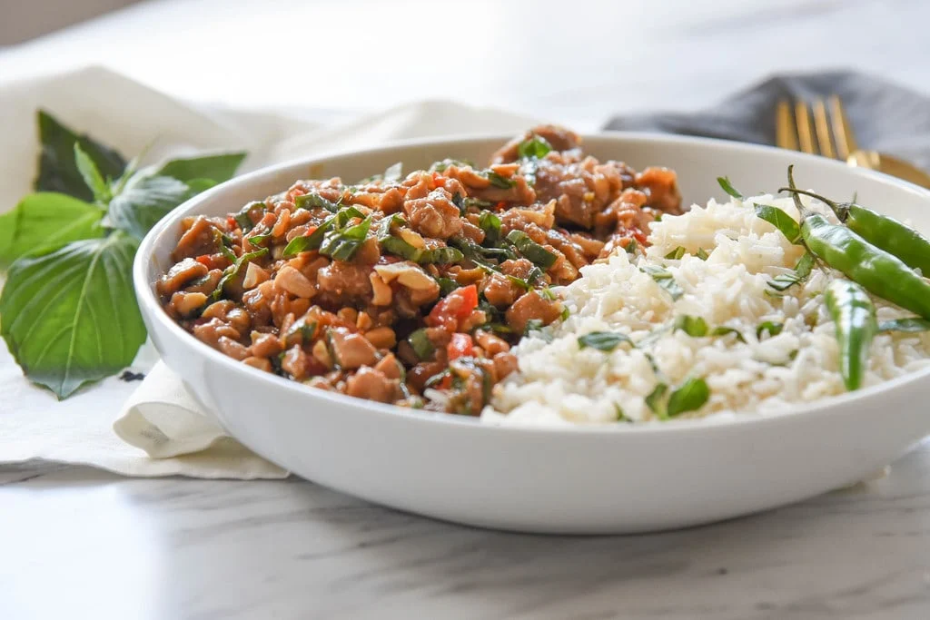 A plated Thai bistro style basil chicken bowl with jasmine rice, glossy chicken and basil, sliced cucumbers, and lime wedges on a warm-toned wooden table