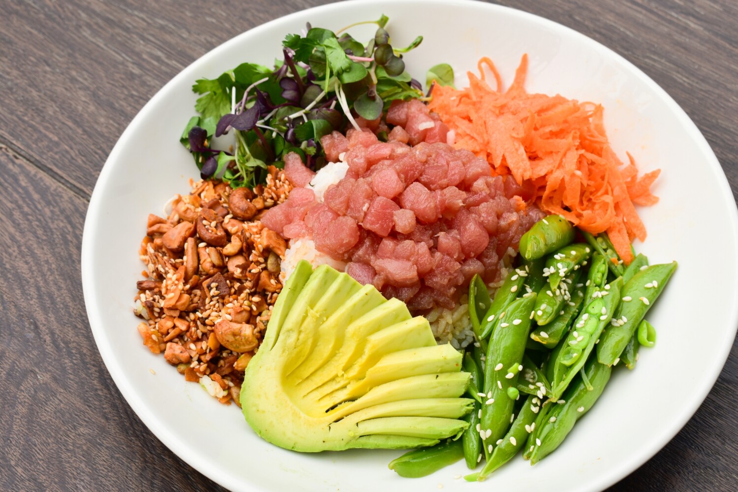 A plated ahi poke bowl with glossy cubes of marinated tuna over sushi rice, topped with sliced avocado, cucumber, scallions, sesame seeds, and a small ramekin of extra sauce on a light wood table