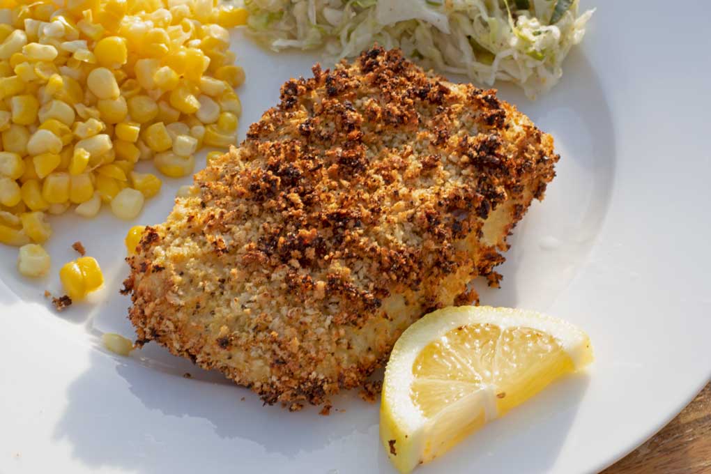 A plated air fryer cod fillet with golden edges, lemon wedges, and chopped parsley on a simple white plate on a warm-toned kitchen table