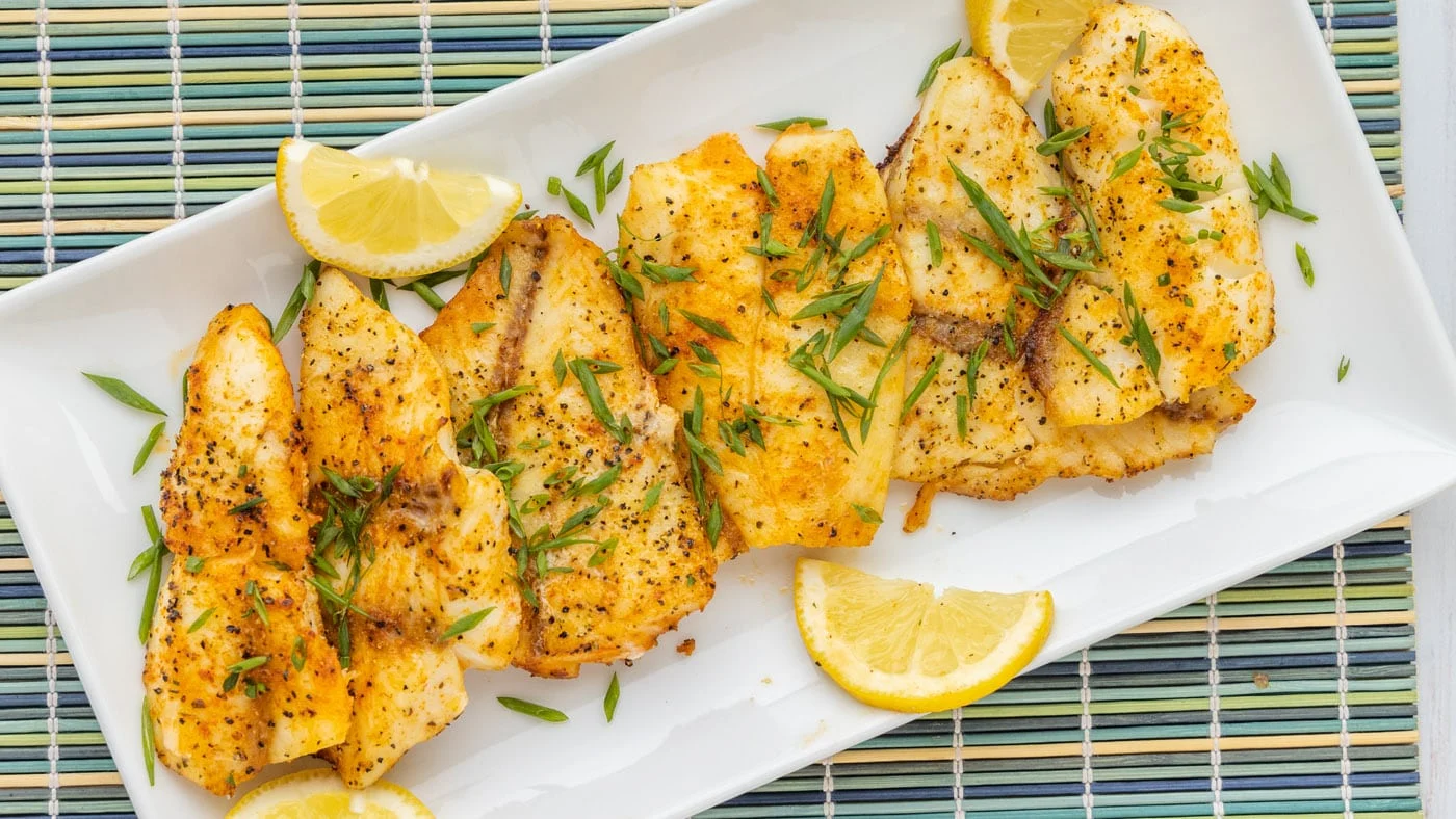 A plated baked orange roughy fillet topped with lemon slices and chopped parsley, with roasted vegetables on the side on a cozy dinner table
