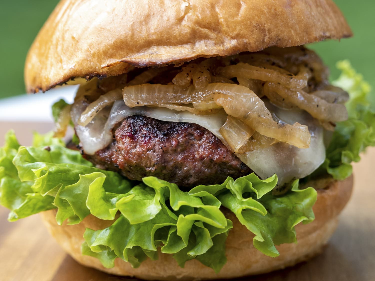 A plated bison cheeseburger on a brioche bun with melted cheddar, pickles, sliced tomato, lettuce, and a smear of tangy sauce on a stoneware plate on a wooden table