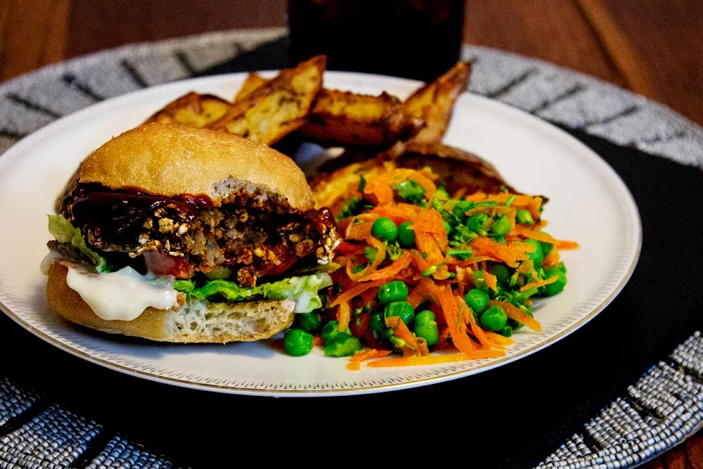 A plated black bean veggie burger on a toasted bun with lettuce, tomato, pickles, and a swipe of sauce, with sweet potato fries on the side on a wooden table in warm natural light