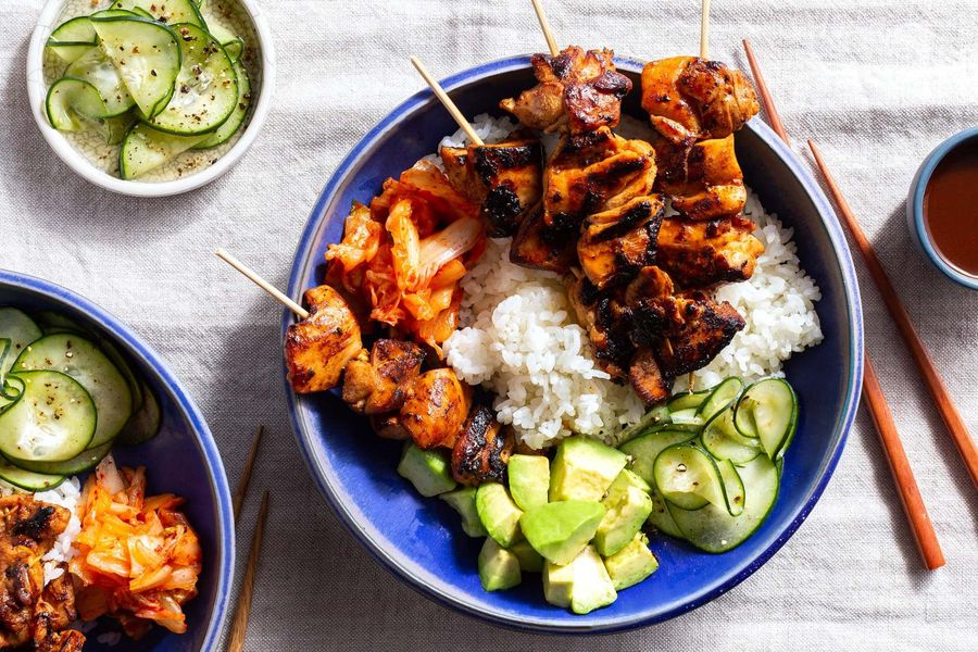 A plated bowl of Korean BBQ chicken thighs with a glossy red-brown glaze, steamed rice, cucumber slices, and sesame seeds on a bright kitchen table