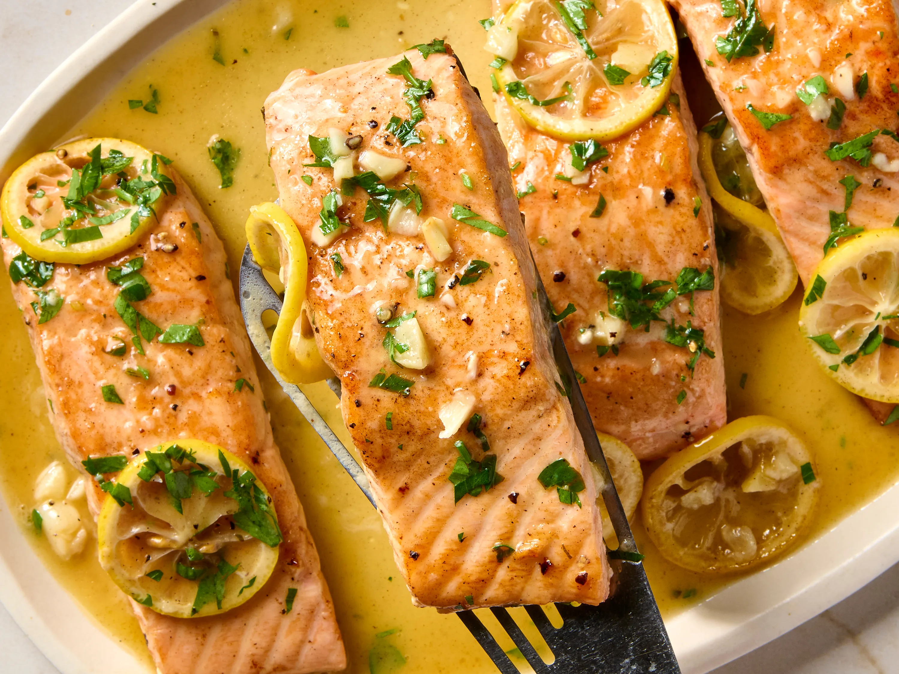 A plated broiled salmon fillet with crisp edges, topped with melted lemon garlic butter and chopped parsley, served with a lemon wedge on a simple white plate on a warm kitchen table