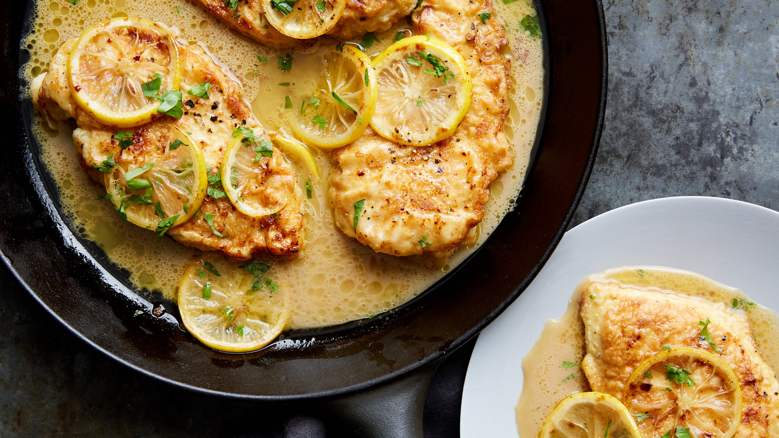 A plated chicken francaise with golden egg-battered chicken cutlets coated in glossy lemon butter sauce, finished with chopped parsley and lemon slices on a neutral dinner plate under warm kitchen lighting
