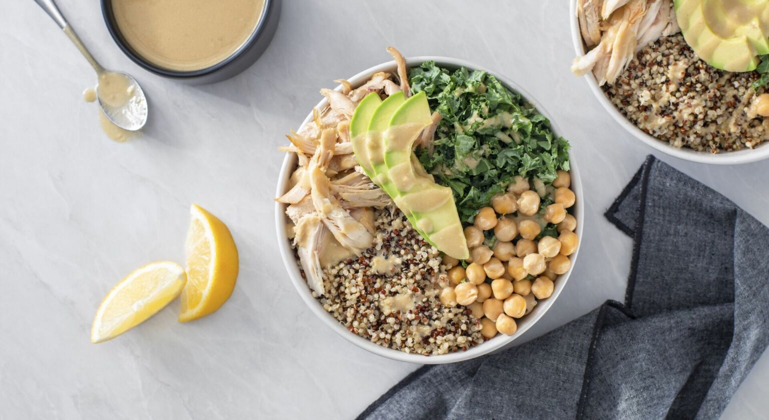 A plated chicken quinoa bowl with sliced lemon-garlic chicken, cucumbers, cherry tomatoes, baby spinach, and a drizzle of lemon tahini sauce on a light kitchen table
