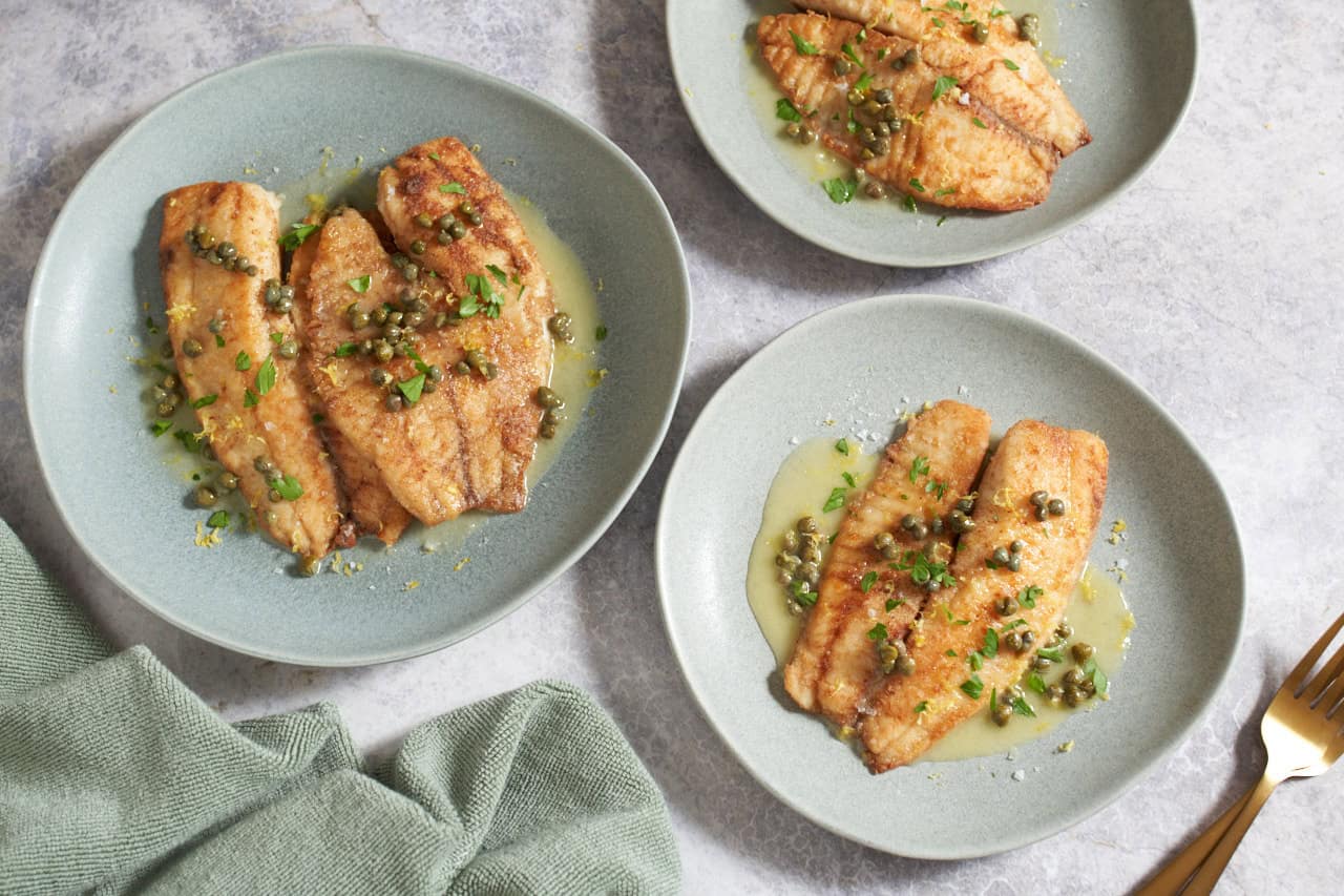 A plated crispy-skinned barramundi fillet topped with glossy lemon-caper butter, served on a white plate with parsley and lemon wedges on a warm wooden table