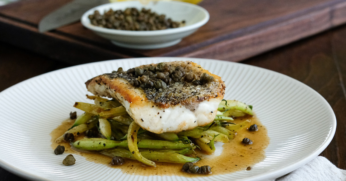 A plated crispy-skinned striped bass fillet topped with lemon-caper pan sauce on a white plate with parsley and lemon wedges, on a warm-toned dinner table