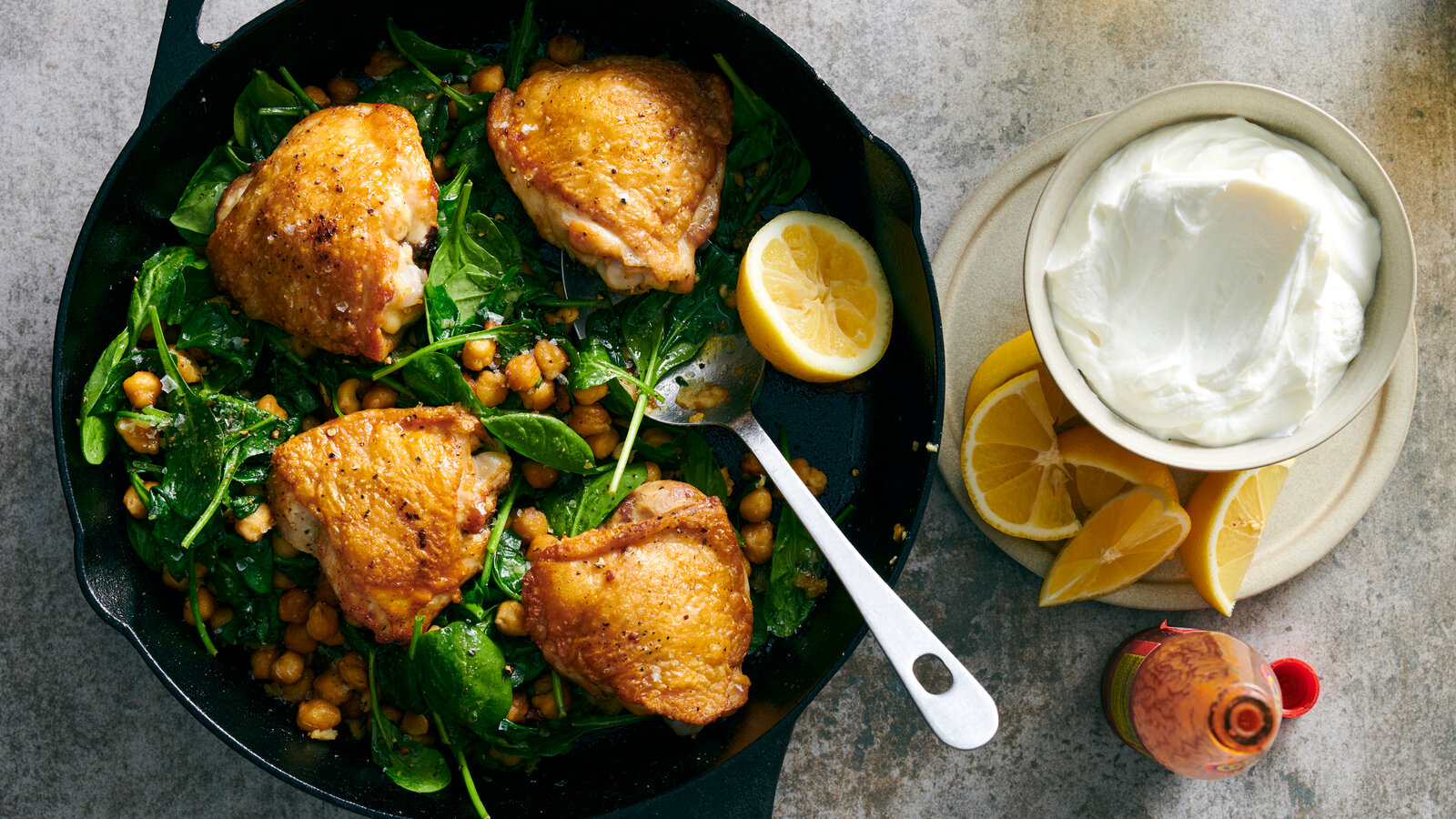 A plated dinner of golden seared chicken thighs topped with crispy spiced chickpeas, served with a swoosh of lemony yogurt sauce and herbs on a ceramic plate on a wooden table
