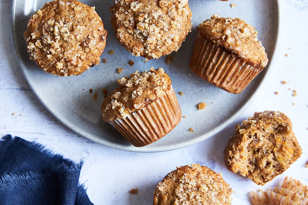 A plated golden carrot muffin on a small breakfast plate with a cup of coffee nearby, shot in bright natural light