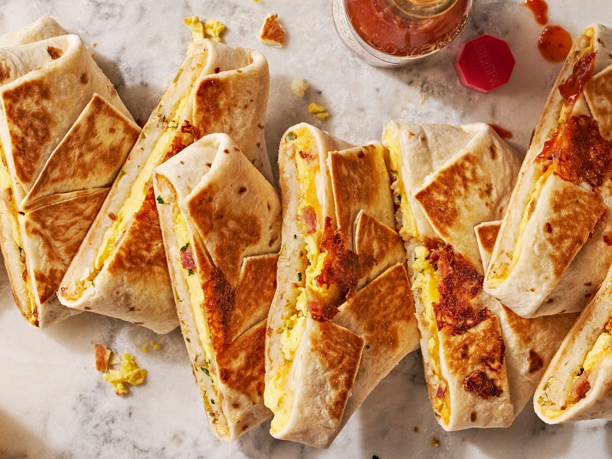 A plated homemade crunchwrap cut in half showing layers of seasoned beef, melted cheese, lettuce, and tomatoes, on a wooden table with a small bowl of sauce and a simple weeknight table setting