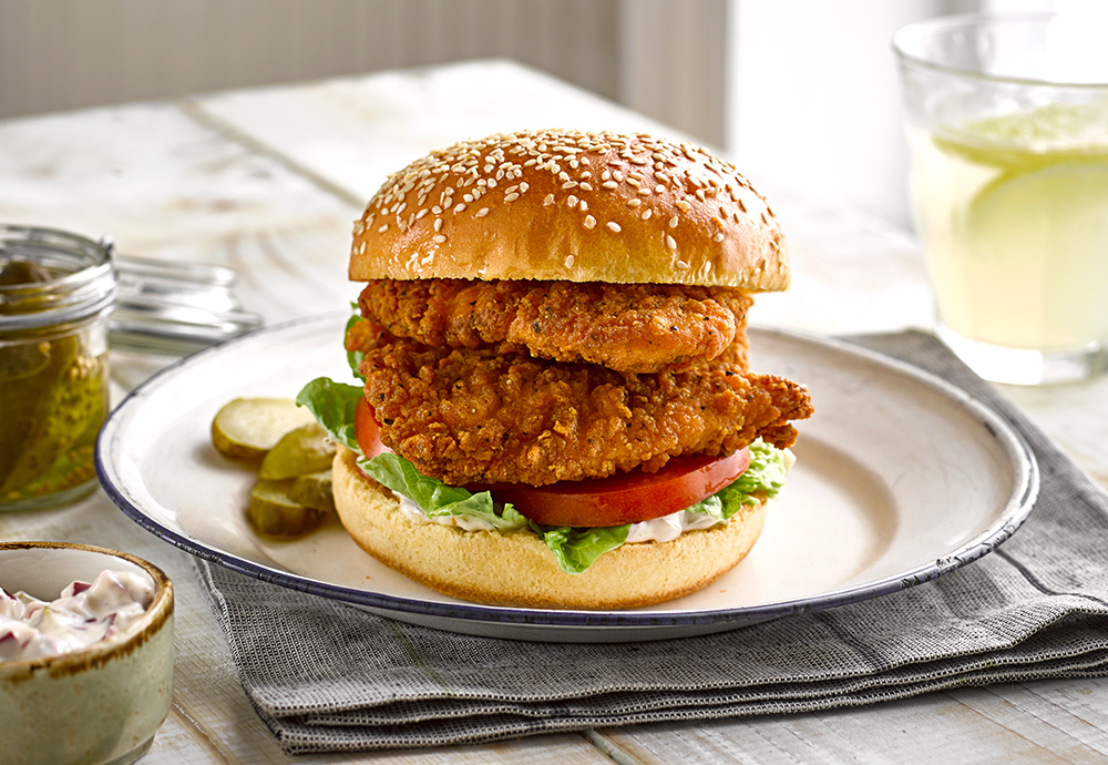 A plated juicy chicken burger on a toasted brioche bun with lettuce, tomato, and a swipe of garlic mayo on a ceramic plate beside a small pile of fries on a wooden kitchen table in warm natural light